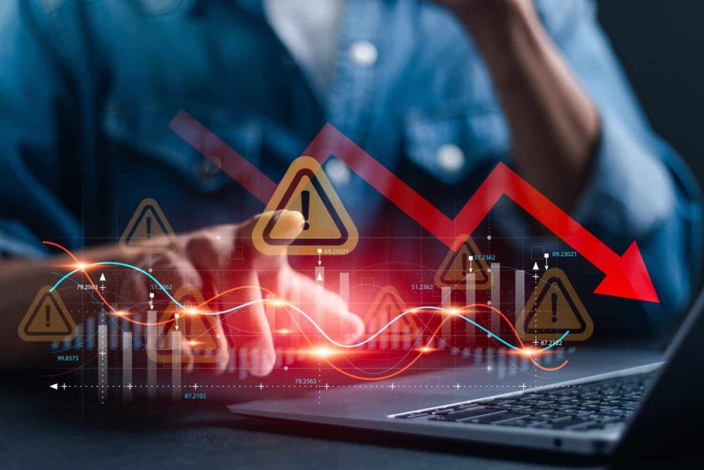A person's hands are shown positioned over a laptop keyboard, illuminated by a warm glow. Overlayed on the scene are various digital financial graphs, including blue and red line charts, bar graphs, and multiple yellow warning triangle symbols. A large, bright red arrow points diagonally downwards from the upper left to the lower right, indicating a significant decline. The overall color scheme is dark with glowing red and blue light effects, suggesting market volatility.