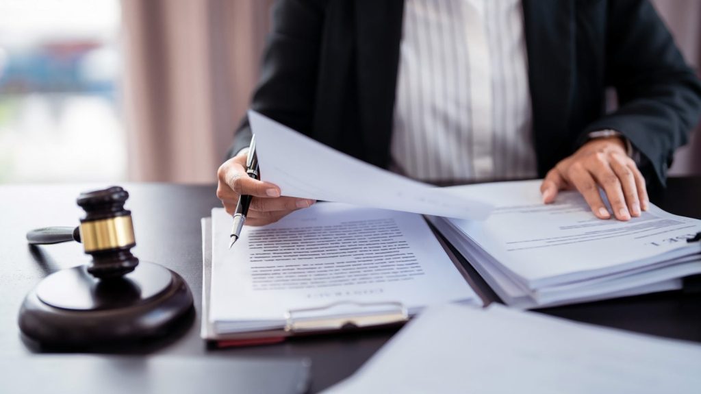 A person in a black suit jacket and white striped shirt sits at a dark desk, holding a black pen over a stack of white documents. Their hands are visible, one holding the pen and the other resting on papers. A wooden gavel with a brass band is placed on the desk to the left of the documents.