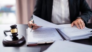 A person in a black suit jacket and white striped shirt sits at a dark desk, holding a black pen over a stack of white documents. Their hands are visible, one holding the pen and the other resting on papers. A wooden gavel with a brass band is placed on the desk to the left of the documents.