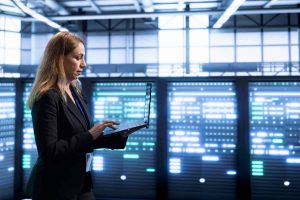 A woman with light brown hair, wearing a black blazer over a blue top, stands in profile to the right, typing on a silver laptop. She is in a dimly lit data center, with numerous server racks filling the background, glowing brightly with blue and white lights.