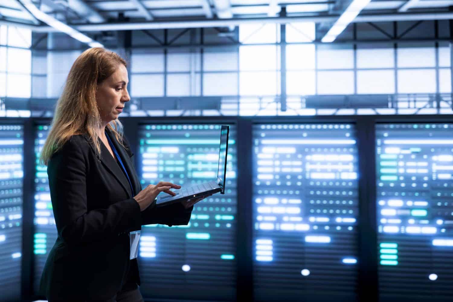 A woman with light brown hair, wearing a black blazer over a blue top, stands in profile to the right, typing on a silver laptop. She is in a dimly lit data center, with numerous server racks filling the background, glowing brightly with blue and white lights.