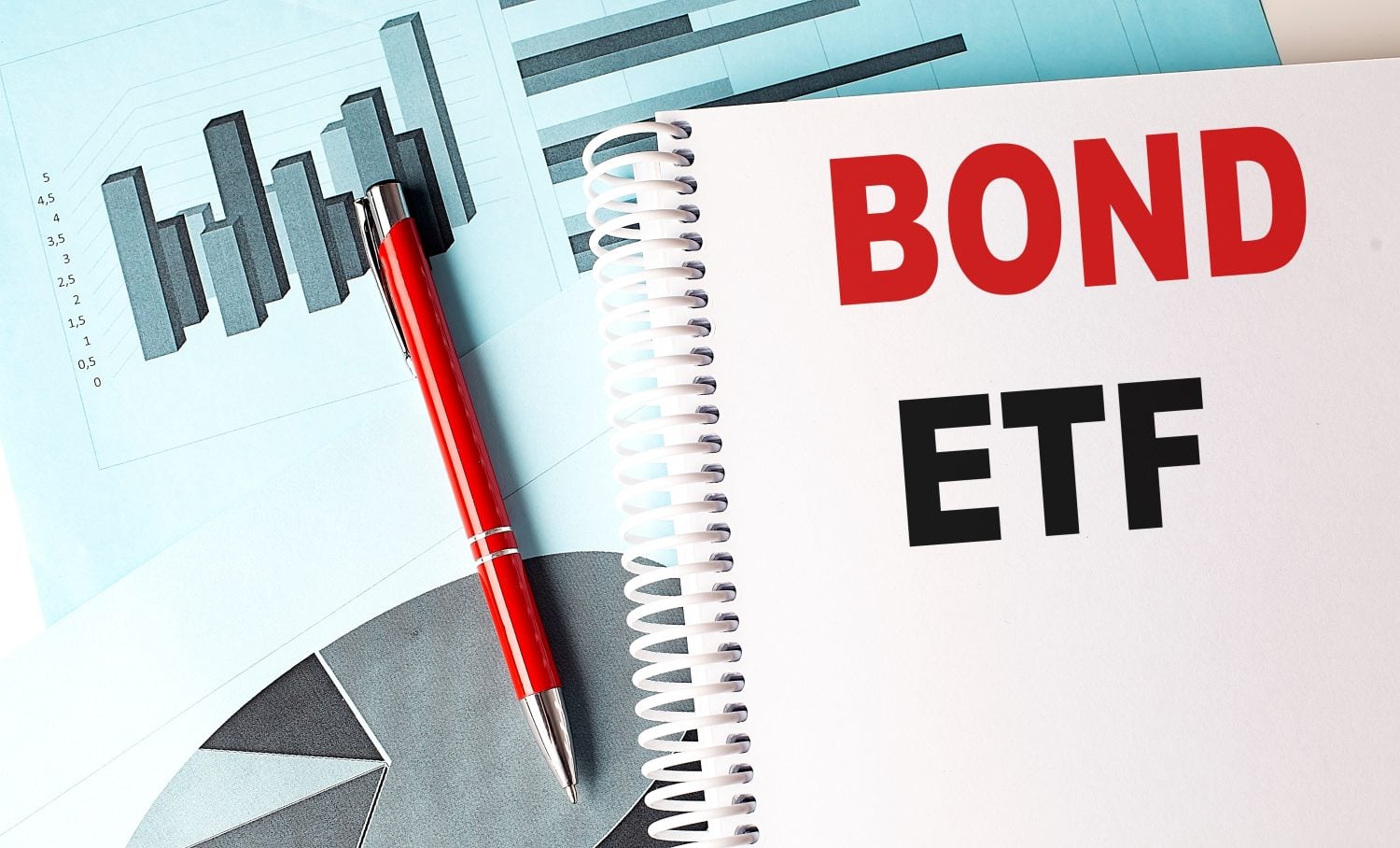 A red pen with a silver tip rests diagonally across a white spiral-bound notebook and a light blue document. The notebook page shows "BOND" in large red letters and "ETF" in large black letters. The document on the left displays a 3D bar chart with numerical labels on the y-axis, and a portion of a grey pie chart is visible at the bottom.