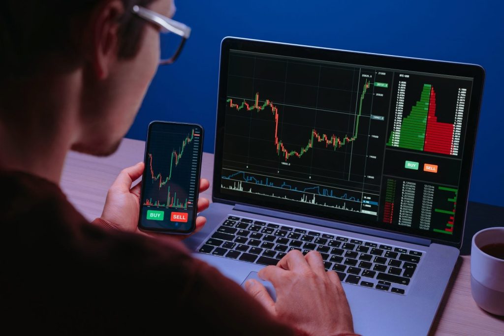 An overhead shot of a person from behind, wearing glasses, intently looking at a laptop and holding a smartphone. Both screens display financial trading charts with green and red candlestick patterns and order book data. The laptop screen shows 'BTC-USD' and price levels, while 'BUY' and 'SELL' buttons are prominently visible on both devices. The person's right hand is positioned on the laptop keyboard, and a white coffee mug sits on the desk to the right.
