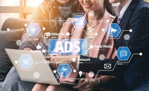 A close-up of three individuals, two women and one person partially obscured, collaboratively looking at a silver laptop screen. A futuristic, glowing blue digital overlay featuring hexagonal icons and text like 'ADS', 'Adwords', 'Social Media', 'Review', 'Network', and symbols for shopping carts, light bulbs, messages, and megaphones is superimposed on the scene. Dotted lines connect these icons to the laptop, suggesting digital advertising and marketing concepts. The background is softly blurred with a warm lens flare.