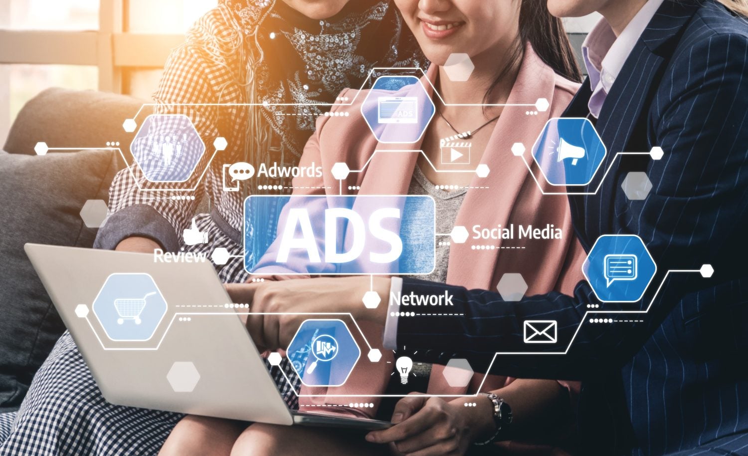 A close-up of three individuals, two women and one person partially obscured, collaboratively looking at a silver laptop screen. A futuristic, glowing blue digital overlay featuring hexagonal icons and text like 'ADS', 'Adwords', 'Social Media', 'Review', 'Network', and symbols for shopping carts, light bulbs, messages, and megaphones is superimposed on the scene. Dotted lines connect these icons to the laptop, suggesting digital advertising and marketing concepts. The background is softly blurred with a warm lens flare.