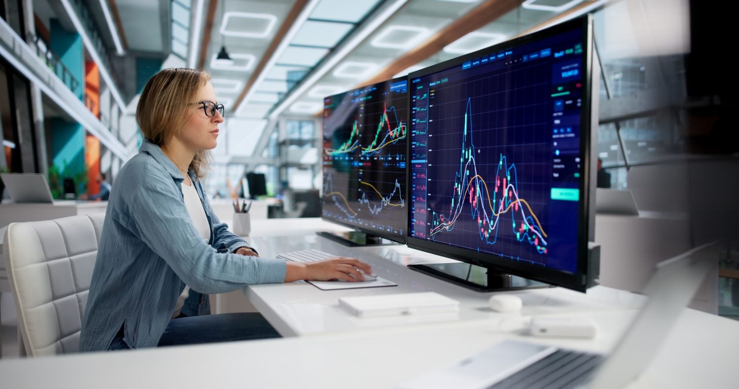 A woman with blonde hair and black-rimmed glasses, wearing a light blue shirt, sits at a white desk in a modern office. She is looking intently at two large computer monitors displaying colorful financial charts and graphs on a dark background. Her right hand rests on a computer mouse. A silver laptop is partially visible on the desk in the foreground to the right, and the bright, spacious office is blurred in the background.