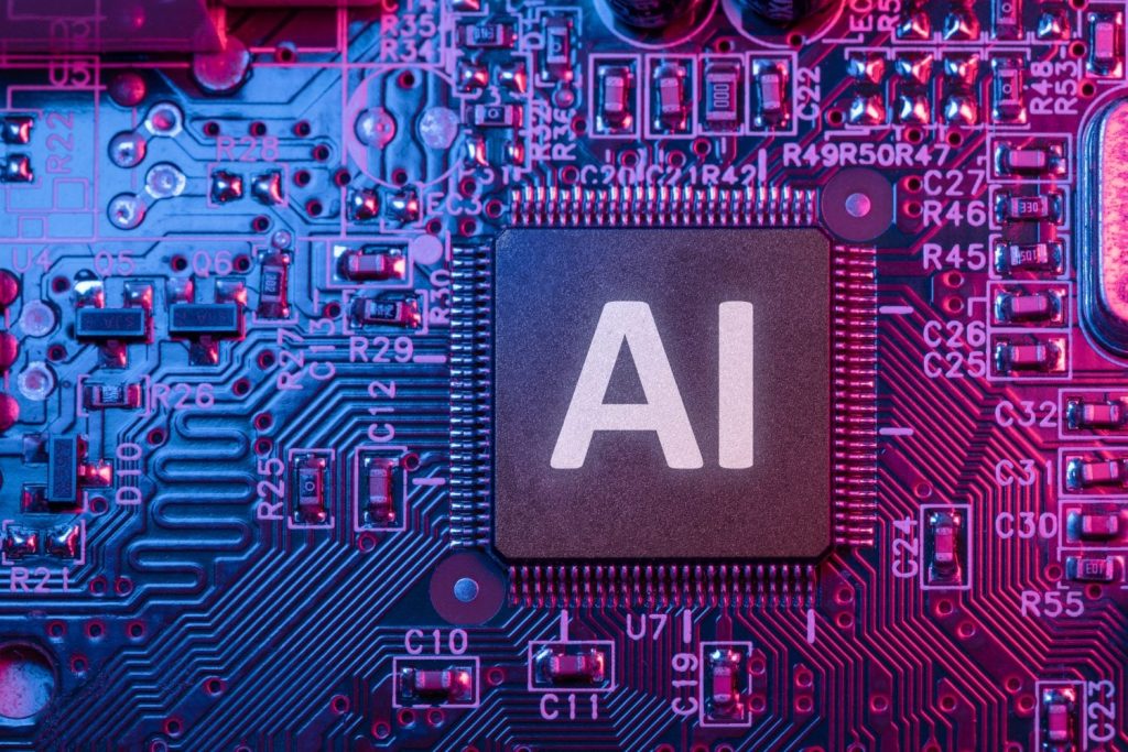 A close-up view of a dark gray microchip with the white letters 'AI' on its surface, centrally positioned on a densely populated electronic circuit board. The board is illuminated with strong blue light on the left and magenta light on the right, casting a colorful glow over the numerous small electronic components, solder points, and etched pathways. Text labels such as R29, C12, U71, R45, and C26 are visible among the components.