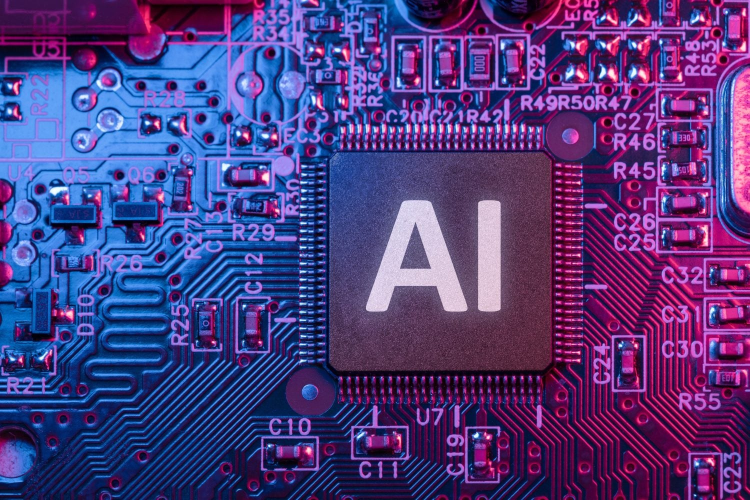 A close-up view of a dark gray microchip with the white letters 'AI' on its surface, centrally positioned on a densely populated electronic circuit board. The board is illuminated with strong blue light on the left and magenta light on the right, casting a colorful glow over the numerous small electronic components, solder points, and etched pathways. Text labels such as R29, C12, U71, R45, and C26 are visible among the components.