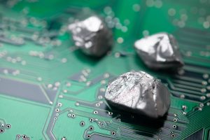 Three rough, silver-grey metal nuggets are placed on a bright green circuit board. The nugget in the foreground is in sharp focus, showing detailed textures, while the two in the background are softly blurred. The circuit board features intricate metallic traces and numerous small, circular solder points.