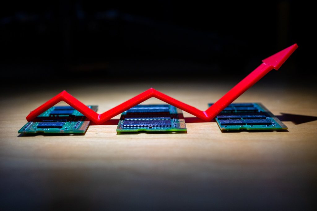 A bright red arrow pointing upwards and to the right, indicating growth or an increase, is placed above and across three green circuit boards, identified as RAM modules. The RAM modules are laid out on a light brown wooden surface, with a dark, blurred background. The arrow appears to be 3D printed and has a segmented, jagged upward path.