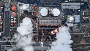 An aerial, high-angle view of a large industrial complex with numerous buildings, storage tanks, and a prominent red and white striped smokestack emitting a thick plume of white smoke. Another large plume of smoke rises from a cylindrical structure in the lower-center left. Multiple circular storage tanks with domed roofs, some dark red and others light gray, are visible throughout the facility. The ground consists of dark paved areas and bare earth, crisscrossed by pipes and industrial equipment under a muted sky.