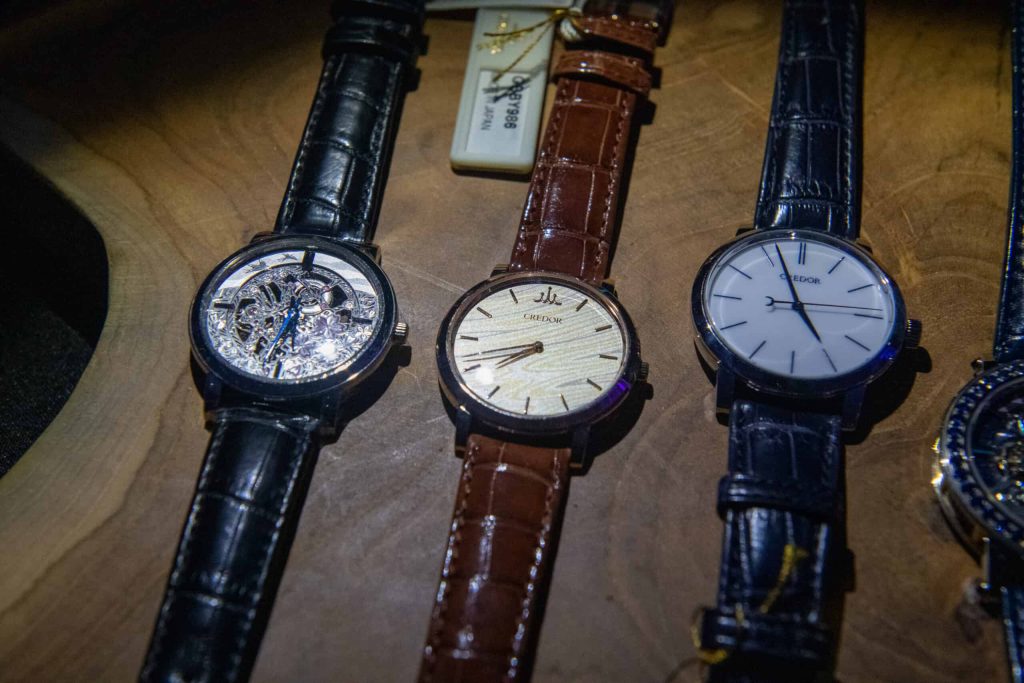 Three luxury watches are displayed on a wooden surface. The leftmost watch has a black crocodile-patterned leather strap and a silver skeletonized dial with intricate gears and blue hands. The middle watch features a brown crocodile-patterned leather strap and a light tan, textured dial with gold hour markers and hands, bearing the brand name 'CREDOR'. The third watch has a black crocodile-patterned leather strap and a clean white dial with black stick hour markers and hands, also labeled 'CREDOR'. A fourth watch is partially visible on the far right.