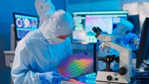 A person dressed in a light blue cleanroom suit, including a head covering, mask, and gloves, holds a large, iridescent silicon wafer with both hands. The wafer reflects a spectrum of colors—pinks, purples, greens, and yellows—from the overhead lights. To the right, a white microscope with multiple objective lenses is visible. In the background, blurred computer monitors display colorful data and graphics, and another person in a cleanroom suit is partially visible. The overall scene is illuminated with a blue light, creating a high-tech and sterile atmosphere.
