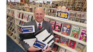 A bald man in a grey suit and red tie smiles directly at the camera while standing in a Blockbuster video store. He holds several stacks of white and black VHS tapes, with the blue 'BLOCKBUSTER VIDEO' logo clearly visible on one of the tapes. Behind him, long rows of white shelves are filled with numerous movie VHS tapes, and blue signs labeled 'KIDS' are prominent in the aisles.