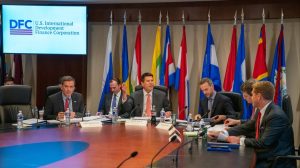 A group of diverse men in business suits are seated around a large, polished oval conference table. In the background, a large screen displays the 'DFC U.S. International Development Finance Corporation' logo. Behind the table, multiple flags from various countries are displayed in a row. Nameplates for 'Amb. Mark Green' and 'Keith Krach' are visible on the table. The men are engaged in discussion, with some looking at documents and others gesturing, amidst water bottles and microphones.