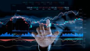 A human hand, with the index finger extended, appears to touch a glowing, translucent blue and red financial chart overlayed on a dark, blurred background. The chart displays candlestick patterns, line graphs, volume indicators, and various numerical values, including '1196.52' prominently. The hand is slightly out of focus in the foreground, while the digital interface shows stock market data and trends in various shades of blue, red, and white on a dark blue-black background.
