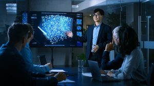 In a modern conference room, an East Asian man wearing glasses and a dark suit stands to the right of a large black monitor, gesturing towards a bright blue, three-dimensional network graph on the screen. He is presenting to several professionals seated at a long, dark table in the foreground, who are attentively listening and looking towards the display. A laptop, papers, and a glass of water are visible on the table.
