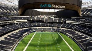 An interior, high-angle view of the empty SoFi Stadium. The bright green football field with white yard lines and numbers is at the center. It is surrounded by tiers of dark blue seats, which are also empty. Above the field, a large, dark, curved overhead structure with the "SoFi Stadium" logo in light blue and white is prominently displayed. The stadium's white roof structure is visible at the top.