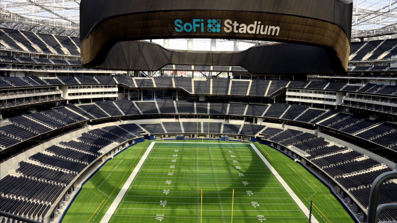 An interior, high-angle view of the empty SoFi Stadium. The bright green football field with white yard lines and numbers is at the center. It is surrounded by tiers of dark blue seats, which are also empty. Above the field, a large, dark, curved overhead structure with the "SoFi Stadium" logo in light blue and white is prominently displayed. The stadium's white roof structure is visible at the top.