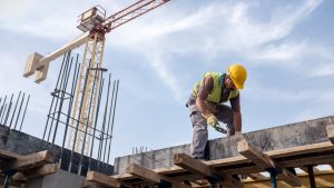 A construction worker wearing a yellow hard hat, lime green safety vest, and gray work pants is bent over, working on a concrete structure with rebar. He holds a tool in his right hand. Wooden planks are part of the formwork below the concrete. In the background, a large white and red construction crane rises against a light blue sky with wispy clouds. The scene is set outdoors on a bright day.