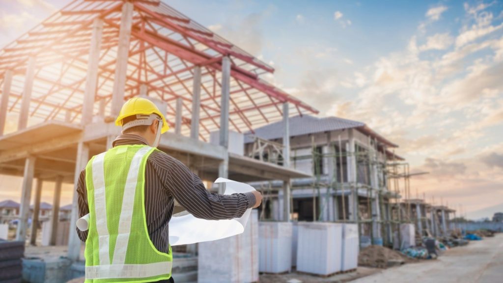 From behind, a construction worker wearing a yellow hard hat and a bright green reflective safety vest holds and reviews large white blueprints. They stand in front of multiple partially constructed modern homes under a bright sky with scattered clouds. The house in the immediate background to the left has a concrete frame, and beyond it, more homes are visible in various stages of construction with scaffolding and exposed frameworks. Sunlight glows warmly from the left side of the frame.