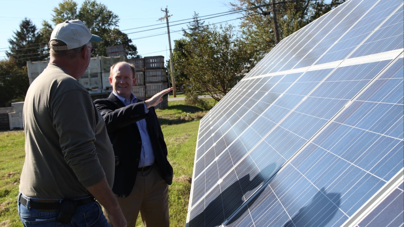 Two men are talking in a grassy field next to a large, angled array of blue solar panels. The man on the left wears a baseball cap and an olive-green shirt, with his back to the viewer. The man on the right, dressed in a dark suit jacket and light blue dress shirt, smiles and gestures with his right hand towards the solar panels. In the background, there are trees, a utility pole with power lines, and stacks of wooden crates under a clear blue sky.