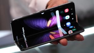 A black foldable smartphone is held partially open in a person's hand, revealing its inner screens. The displays feature a vibrant purple and orange butterfly wing wallpaper, with app icons and a 'Say Hey Google' search bar visible on the right panel. The phone is angled slightly, highlighting its innovative hinge design.