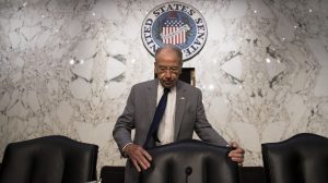 An older man with glasses, wearing a gray suit, white shirt, and navy tie, looks down pensively while holding the back of a black leather chair. Behind him is a white marble wall with dark gray veining, featuring a large, circular United States Senate seal.