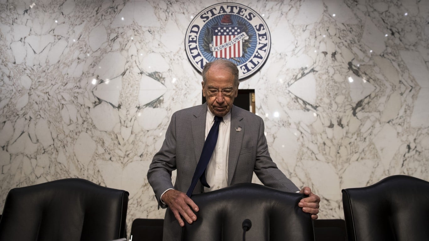 An older man with glasses, wearing a gray suit, white shirt, and navy tie, looks down pensively while holding the back of a black leather chair. Behind him is a white marble wall with dark gray veining, featuring a large, circular United States Senate seal.
