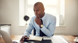 A dark-skinned man with a shaved head, wearing a light blue dress shirt and a dark tie, sits at a wooden desk. He looks intently at a silver laptop on the left, with his right hand resting on his chin in a thoughtful pose. His left hand holds a pen above an open white notebook. A gold watch is visible on his left wrist. In the background, a sunny window, a dark globe, and white folders on a shelf are visible. A white coffee cup is on the far right.