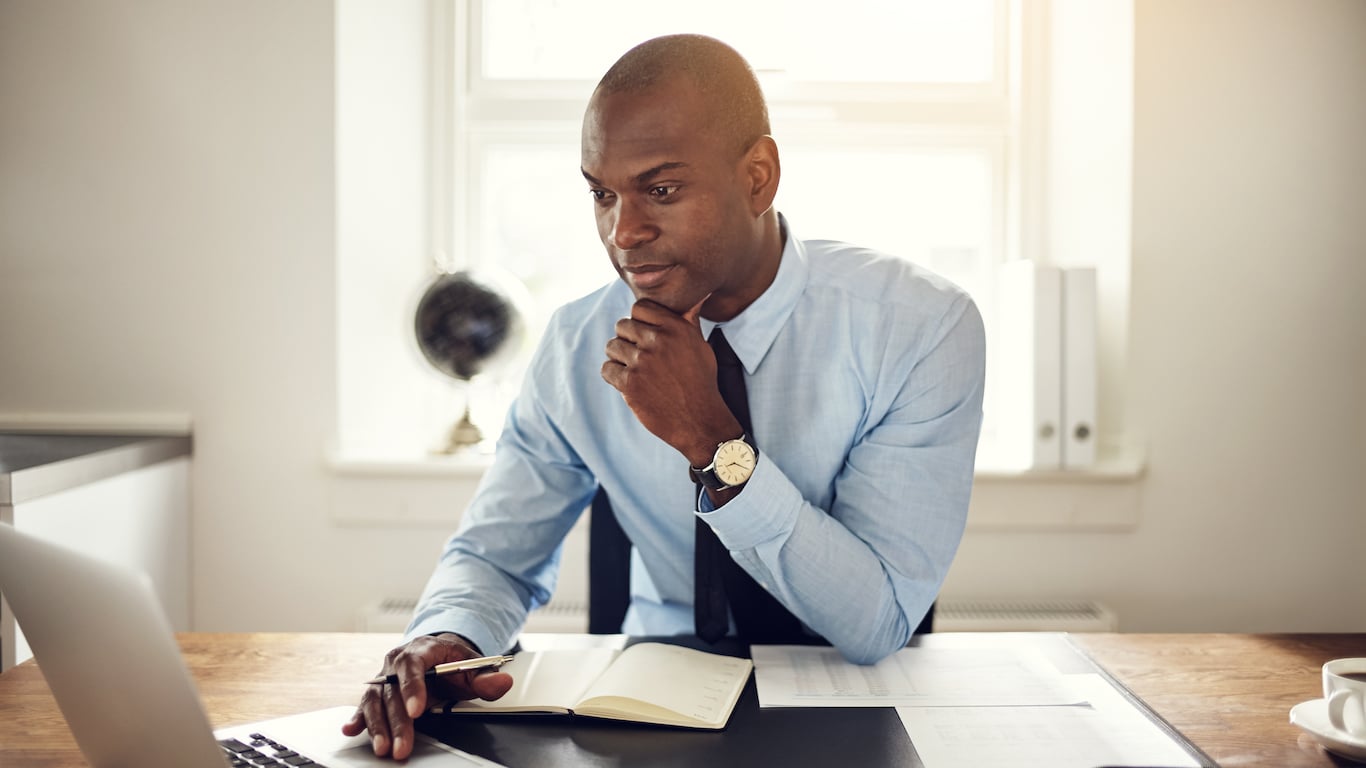 A dark-skinned man with a shaved head, wearing a light blue dress shirt and a dark tie, sits at a wooden desk. He looks intently at a silver laptop on the left, with his right hand resting on his chin in a thoughtful pose. His left hand holds a pen above an open white notebook. A gold watch is visible on his left wrist. In the background, a sunny window, a dark globe, and white folders on a shelf are visible. A white coffee cup is on the far right.