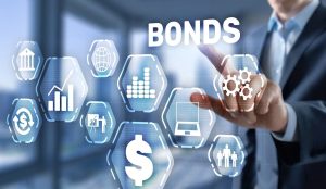 A person in a business suit points a finger towards a glowing hexagonal icon containing three gears, part of a network of white outlined financial symbols, against a blurred blue and white background. The prominent white word 'BONDS' appears in the upper right. Other hexagonal icons include a bank building, a globe, multiple bar charts, a grid of dots, a laptop, a dollar sign, and a group of three people.