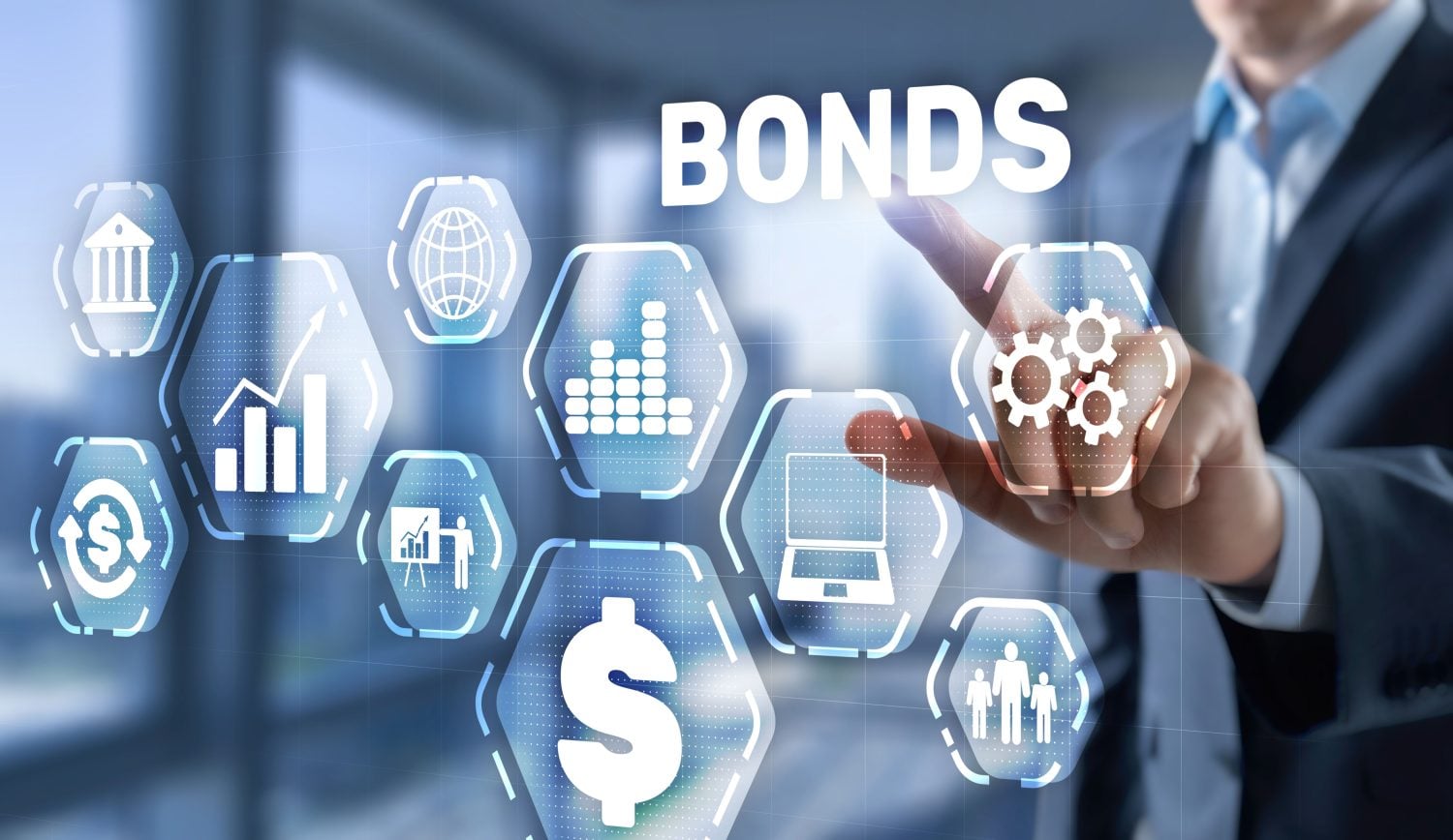 A person in a business suit points a finger towards a glowing hexagonal icon containing three gears, part of a network of white outlined financial symbols, against a blurred blue and white background. The prominent white word 'BONDS' appears in the upper right. Other hexagonal icons include a bank building, a globe, multiple bar charts, a grid of dots, a laptop, a dollar sign, and a group of three people.