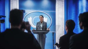 A woman with dark hair pulled back, wearing a gray suit jacket and a light blue collared shirt, stands at a dark gray podium. Her hands are open and gesturing outwards. Behind her is a vibrant blue wall with a large circular emblem featuring a globe and laurel wreaths. In the blurred foreground, the backs of several people, possibly members of the press or an audience, are visible, holding cameras or listening.