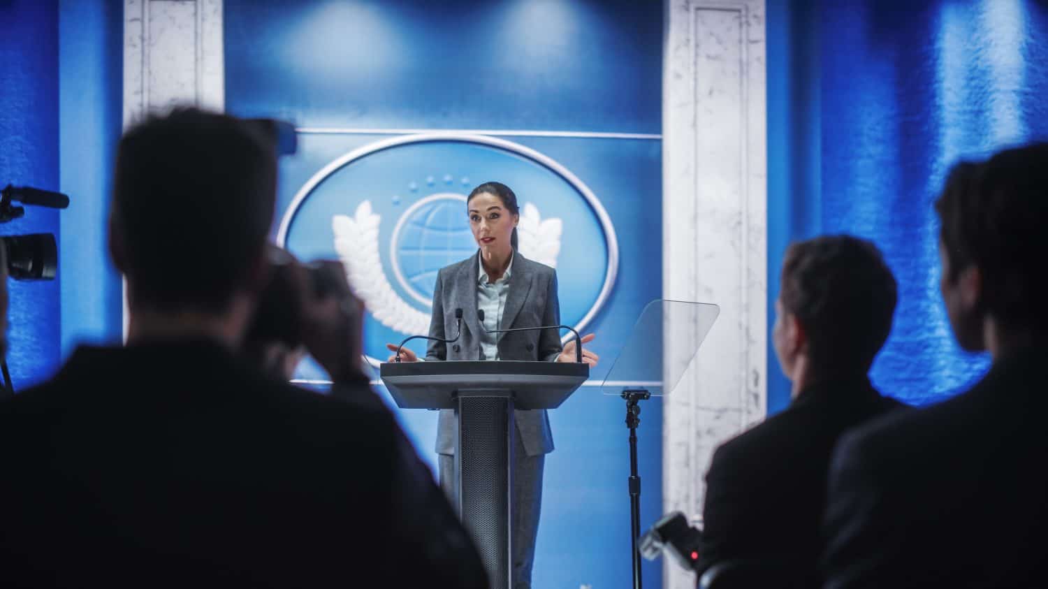 A woman with dark hair pulled back, wearing a gray suit jacket and a light blue collared shirt, stands at a dark gray podium. Her hands are open and gesturing outwards. Behind her is a vibrant blue wall with a large circular emblem featuring a globe and laurel wreaths. In the blurred foreground, the backs of several people, possibly members of the press or an audience, are visible, holding cameras or listening.
