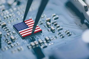 A close-up shot shows a small, rectangular microchip with the design of the American flag on its surface. A pair of silver tweezers is holding the chip, positioning it over a blue circuit board filled with various small electronic components and intricate traces. The foreground and background are slightly blurred, emphasizing the chip and tweezers.