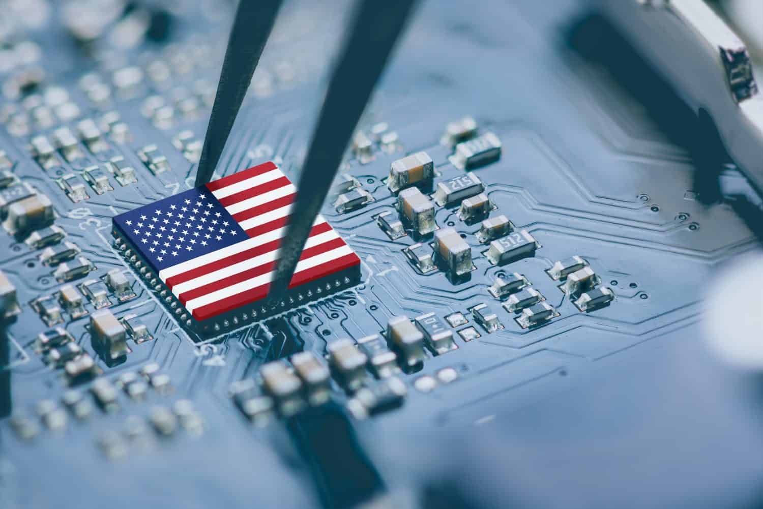A close-up shot shows a small, rectangular microchip with the design of the American flag on its surface. A pair of silver tweezers is holding the chip, positioning it over a blue circuit board filled with various small electronic components and intricate traces. The foreground and background are slightly blurred, emphasizing the chip and tweezers.