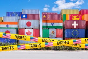 Tall stacks of colorful shipping containers, each adorned with the national flag of a different country such as the EU, Taiwan, Japan, Canada, India, China, Mexico, Sri Lanka, and Switzerland. The foreground is crossed by yellow caution tape, prominently displaying the word 'TARIFFS' in black letters and small US flags, creating a visual barrier. A bright blue sky with sparse clouds forms the background.