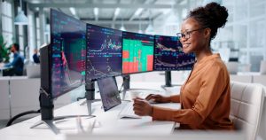 A smiling Black woman with her hair in a bun and wearing glasses sits at a desk, looking at three large computer monitors displaying colorful financial charts and graphs. She is holding a computer mouse with her right hand and a keyboard is in front of her. A tablet showing similar data and a white coffee cup are also on the white desk. The background shows a blurred modern office environment with other people working.