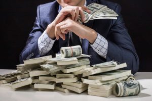 Close-up of a person in a dark blue suit and a white-and-blue checkered shirt, with their hands clasped in front of their face, sitting at a table piled high with stacks and rolls of US hundred-dollar bills. The background is dark and out of focus.