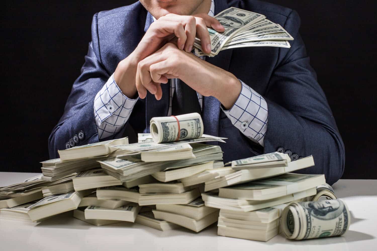 Close-up of a person in a dark blue suit and a white-and-blue checkered shirt, with their hands clasped in front of their face, sitting at a table piled high with stacks and rolls of US hundred-dollar bills. The background is dark and out of focus.