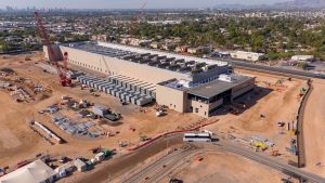 An aerial view of a large construction site featuring a long, light-brown colored data center building with numerous grey rectangular cooling units on its roof and along one side. A very tall red crane stands prominently near the building. The surrounding ground is reddish-brown dirt, covered with construction materials, smaller buildings, equipment, and vehicles. In the background, there are residential areas, lush green trees, a distant city skyline, and mountains under a bright sky.