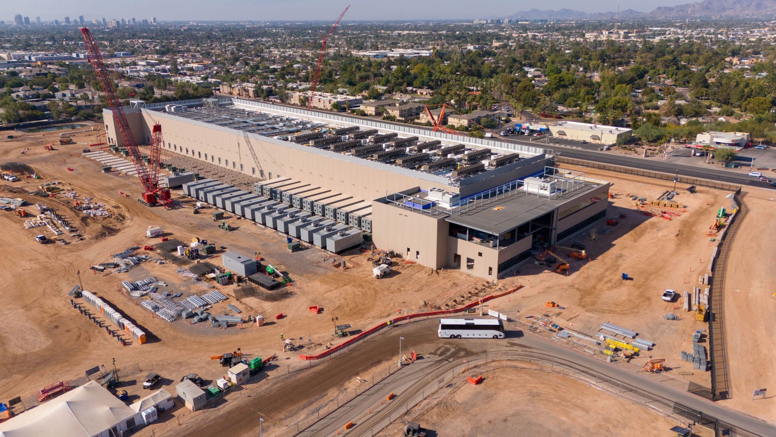 An aerial view of a large construction site featuring a long, light-brown colored data center building with numerous grey rectangular cooling units on its roof and along one side. A very tall red crane stands prominently near the building. The surrounding ground is reddish-brown dirt, covered with construction materials, smaller buildings, equipment, and vehicles. In the background, there are residential areas, lush green trees, a distant city skyline, and mountains under a bright sky.