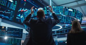 Two financial professionals, seen from behind, are in a dimly lit trading room filled with numerous glowing blue and green computer screens displaying stock charts and financial data. The man in the foreground has both arms raised in a gesture of reaction, while a woman stands nearby. The screens show various graphs and numbers, some with red and green indicators, creating an intense, data-rich environment.