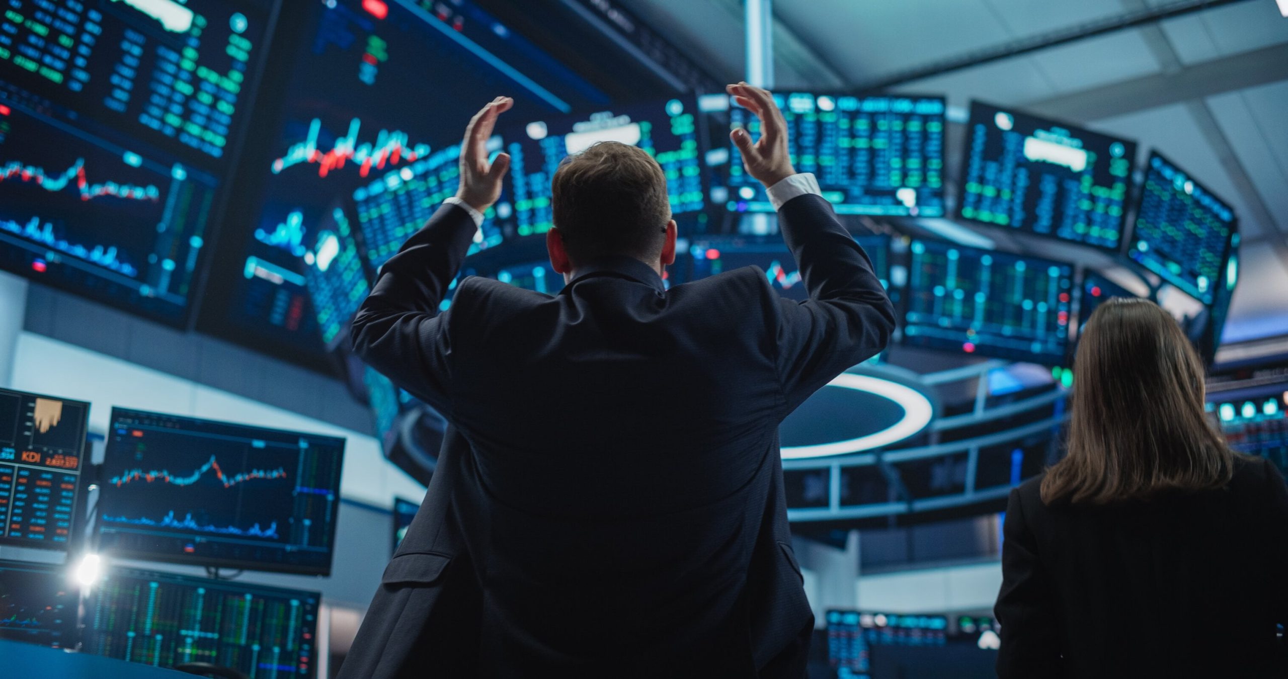 Two financial professionals, seen from behind, are in a dimly lit trading room filled with numerous glowing blue and green computer screens displaying stock charts and financial data. The man in the foreground has both arms raised in a gesture of reaction, while a woman stands nearby. The screens show various graphs and numbers, some with red and green indicators, creating an intense, data-rich environment.