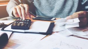 A close-up overhead shot shows a person's hands engaged in financial work on a desk. One hand operates a black calculator with red and white buttons, while the other holds a stack of white paper receipts. An open spiral notebook, a black smartphone, and various financial documents with charts and graphs are spread across the wooden desk. The person wears a grey long-sleeved shirt, with warm sunlight illuminating the scene from the left.