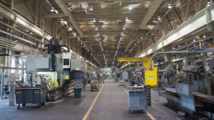 A wide-angle interior view of a brightly lit industrial factory floor. Large, grey metal manufacturing machines line both sides of a central aisle, which has yellow dividing lines on its dark grey surface. The ceiling is high, revealing exposed metal beams, ductwork, and numerous overhead fluorescent lights. Several rolling carts and bins are scattered around the equipment, suggesting ongoing operations. The overall impression is one of large-scale, heavy industry.