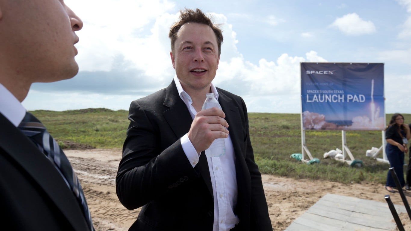 A medium shot shows Elon Musk, a white man with short brown hair, wearing a black suit and white shirt, holding a clear water bottle in his right hand. He is looking slightly to his right and smiling faintly. To his left, the profile of another man in a suit is visible. In the background, a large dark blue banner with "SPACEX" and "SPACEX'S SOUTH TEXAS LAUNCH PAD" is visible, featuring a graphic of a rocket launch. The setting is an outdoor, open field with green grass and some dirt paths under a partly cloudy blue sky.