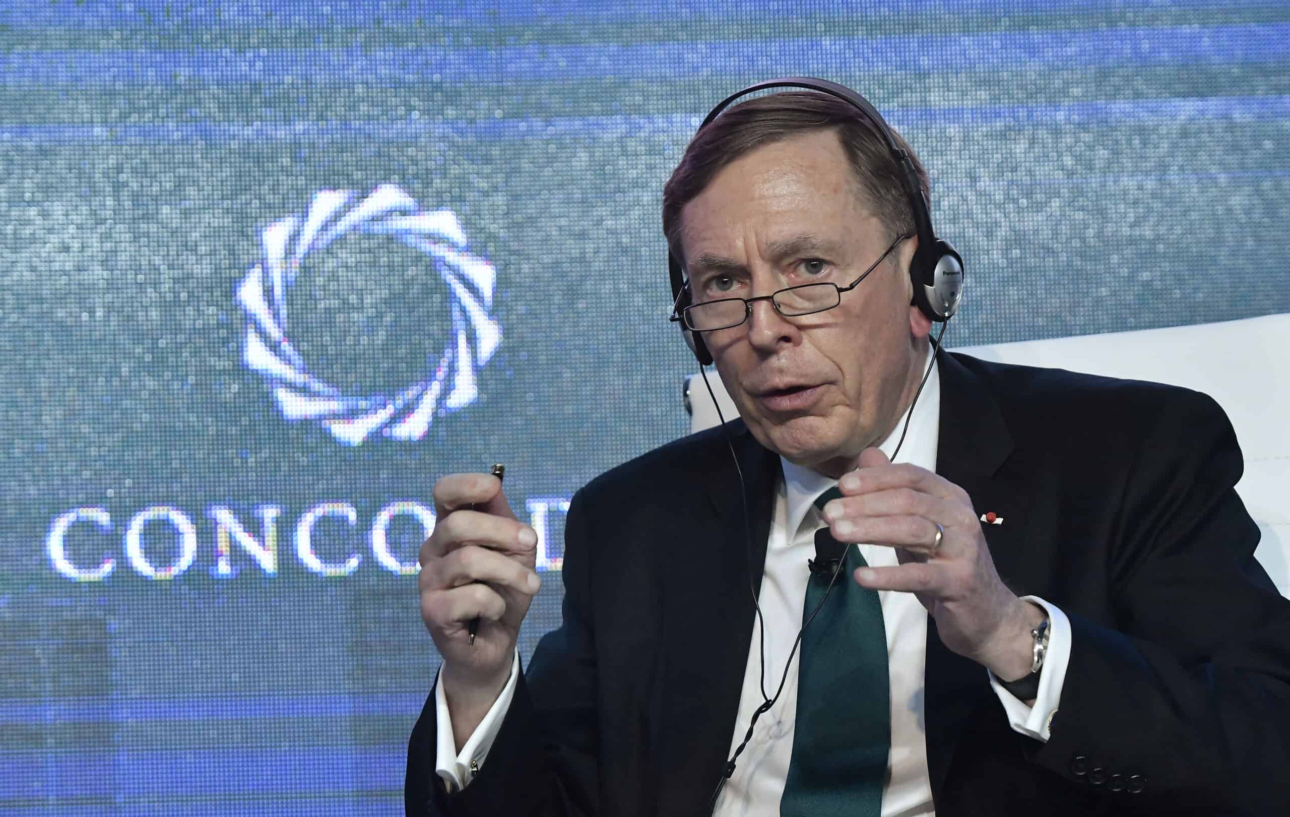 A man with short brown hair, wearing glasses, black headphones, a dark suit, white shirt, and a dark green tie, speaks animatedly, holding a pen in his right hand and gesturing with his left. He is seated in front of a blue screen displaying the partially visible white logo and text 'CONCORDIA'.