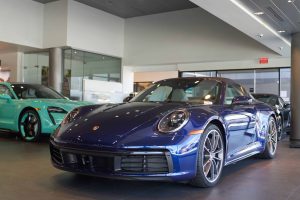 A dark blue Porsche 911 sports car is prominently featured in the foreground of a brightly lit dealership showroom. Behind it, a light teal Porsche Taycan electric sedan is visible to the left, and another dark car is partially seen further back on the right. The showroom has reflective floors and glass walls, creating a sleek, modern environment.